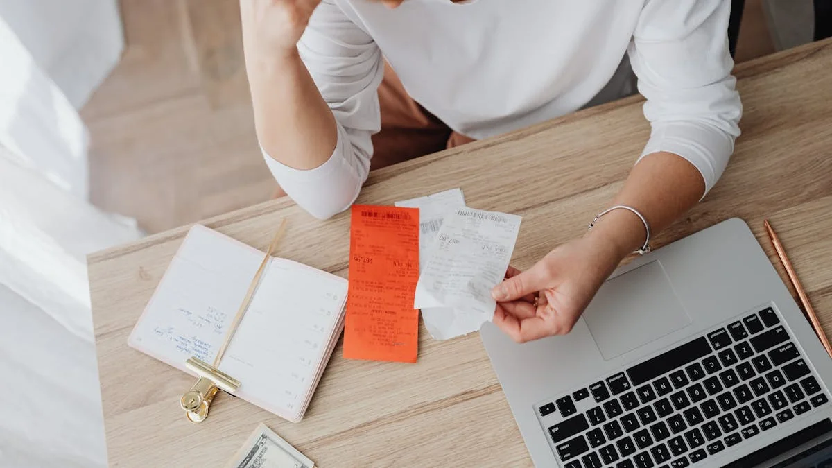 A person reviews their software subscriptions on a laptop, a pile of monthly bills beside them