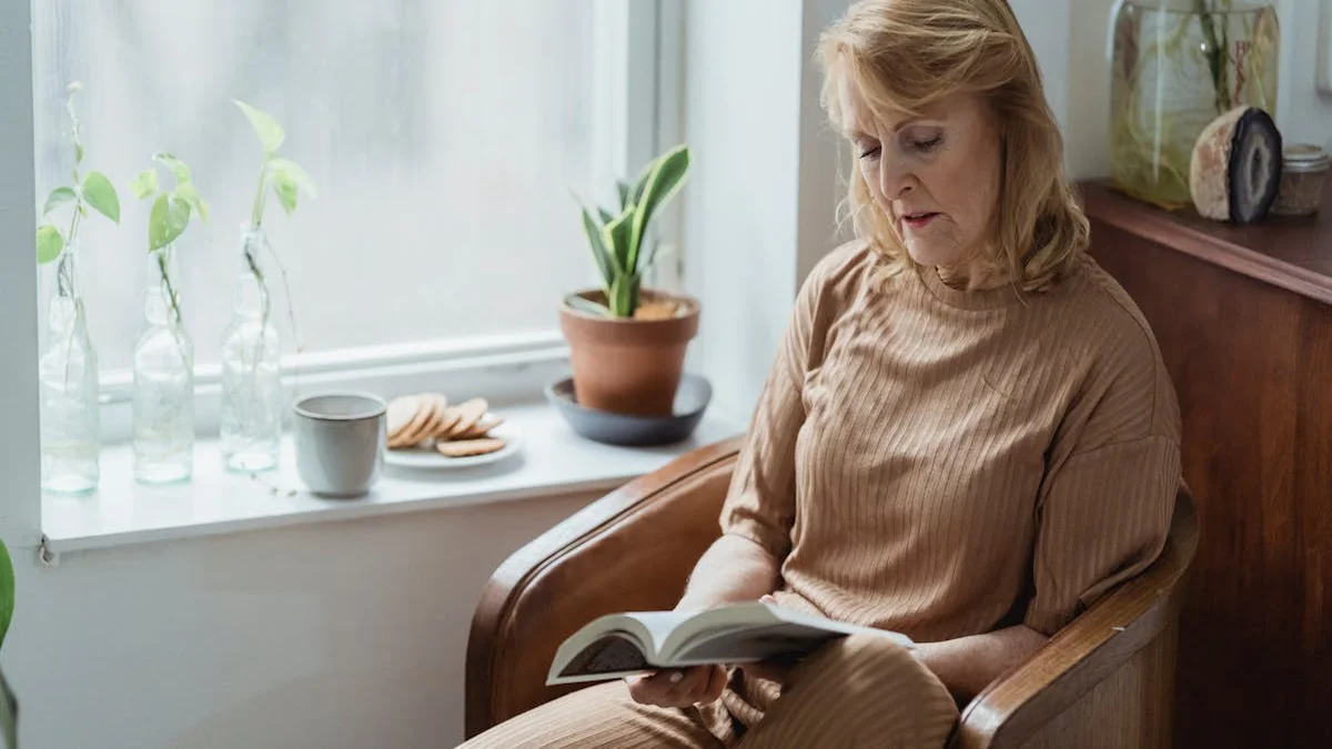A senior woman relaxing in a sunlit armchair, holding a mug of tea and looking thoughtfully at a photo album